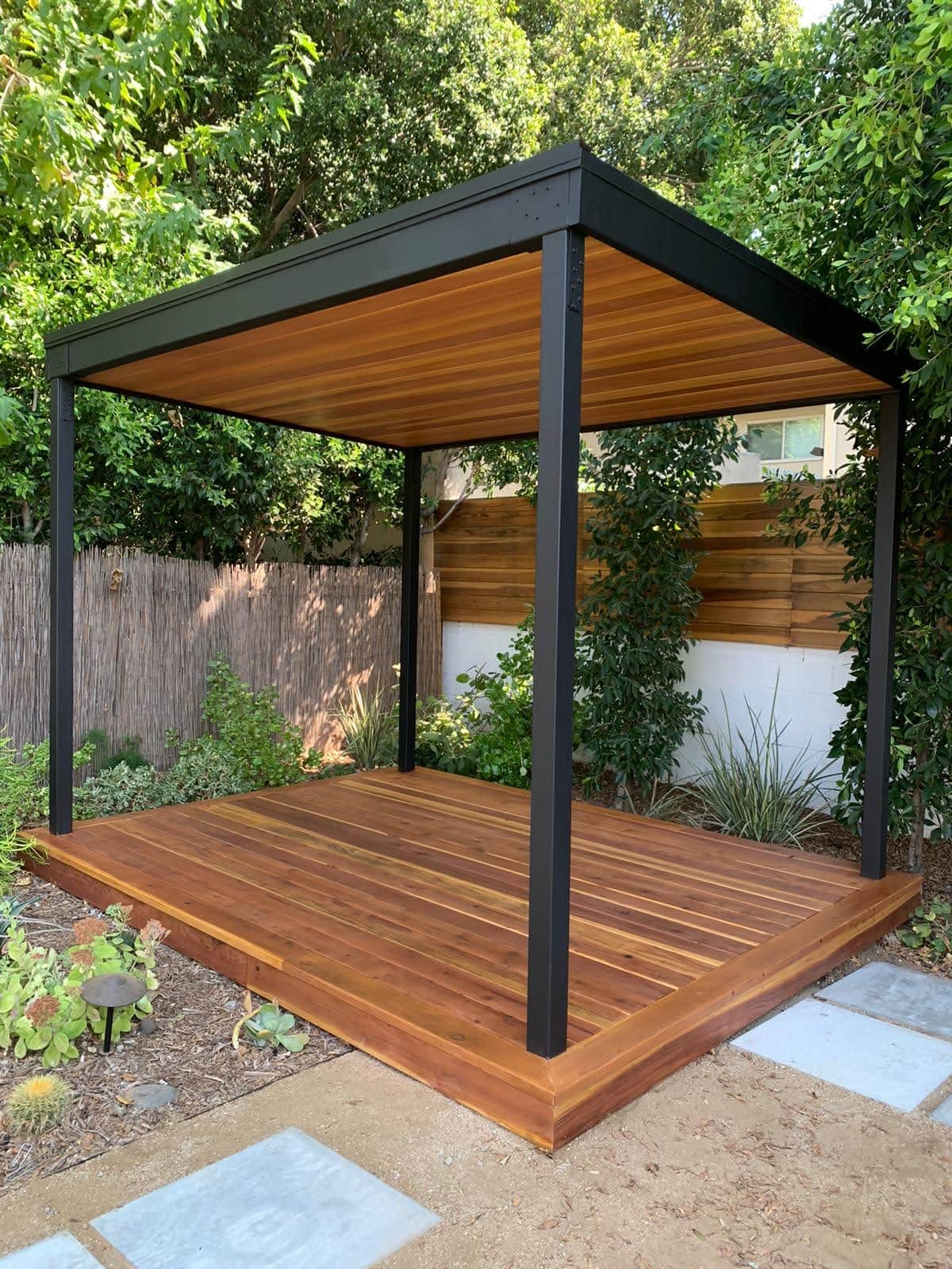 Outdoor deck with black pergola for shade in a lush garden setting.